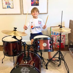 This image shows George, a happy young boy, smiling and sitting behind his drum kit, drumsticks in hand.